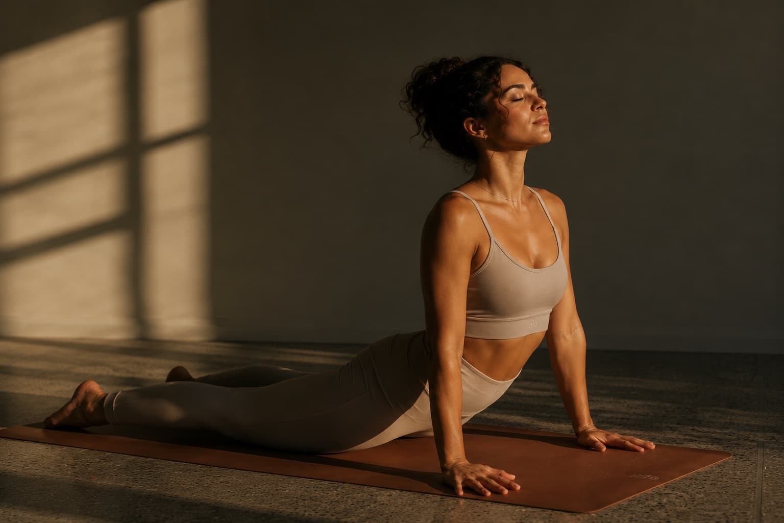 A woman in an upward-facing dog yoga pose on a mat, eyes closed, lit by warm sunlight